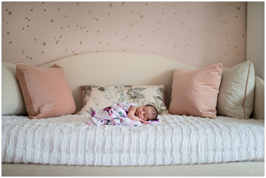 Newborn girl is lying on her daybed surrounded by pink pillows and in a pink wrap