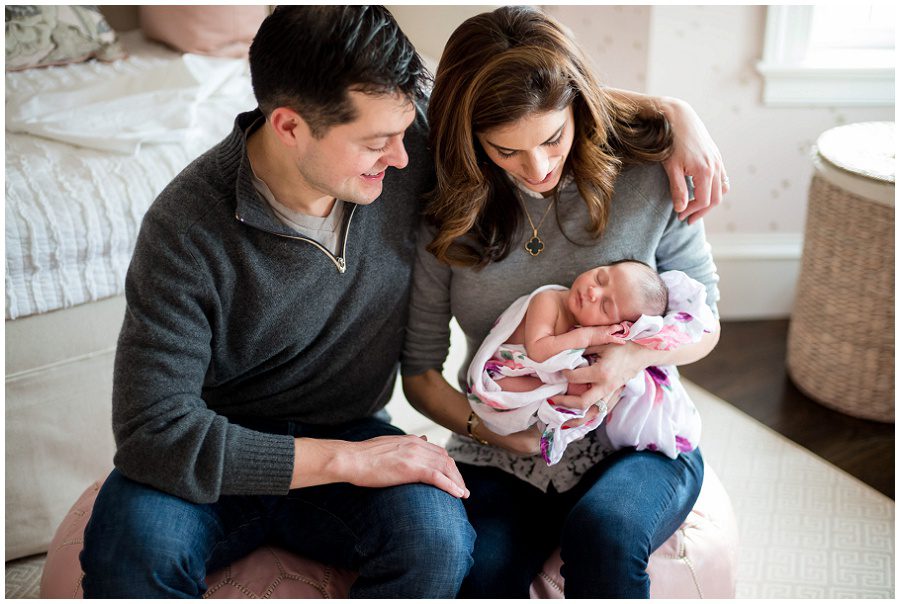 mother holding baby in home session on pink backless chairs vertical to parents holding sleeping newborn swaddled in blanket being held by father