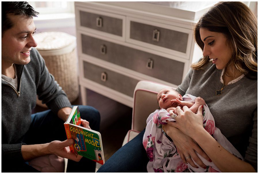 mother holding baby in home session on pink backless chairs vertical to parents holding sleeping newborn swaddled in blanket listening to father reading a book