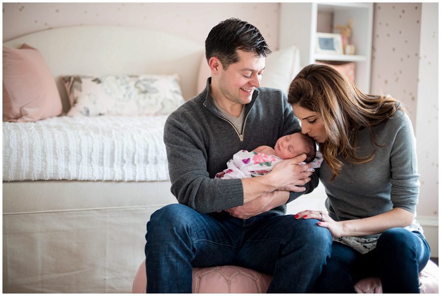 Father holding baby in home session on pink backless chairs vertical to parents holding sleeping newborn swaddled in blanket and mom kissing head of baby