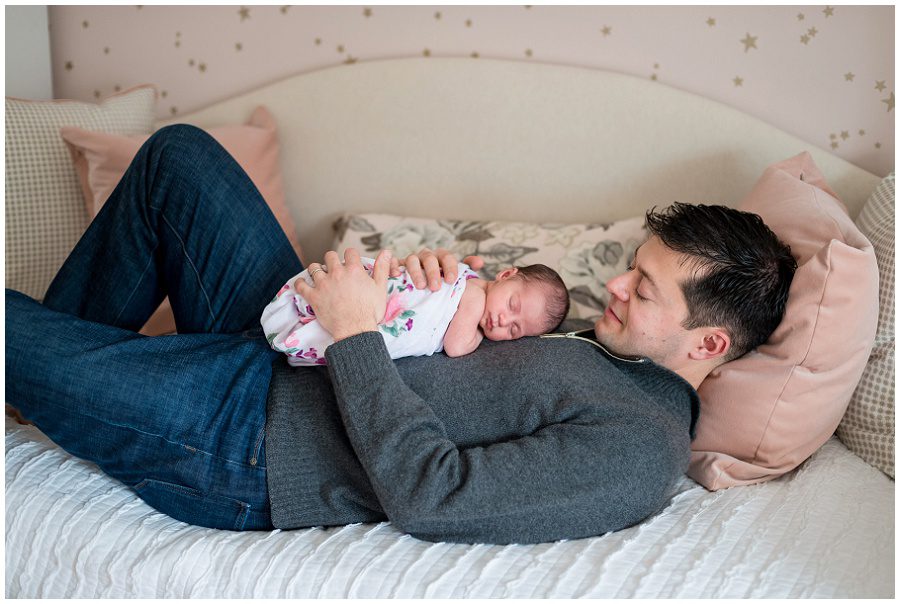 Father lying on day bed with newborn on chest as he looks down at baby that is swaddled in a white wrap