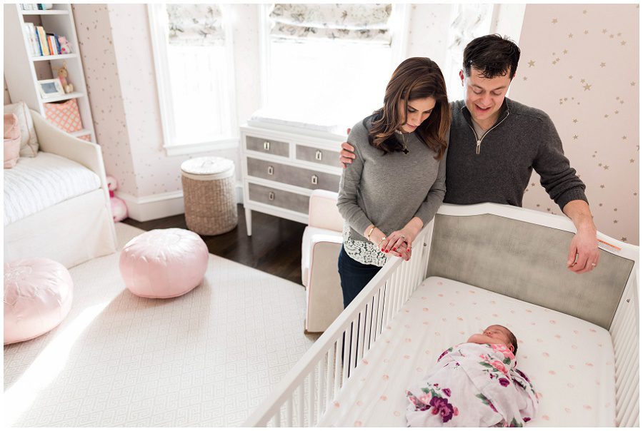 parents looking at newborn as she lies in her crib swaddled in a blanket