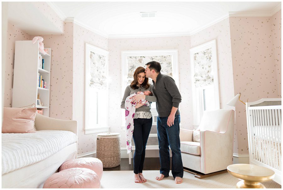 Father kisses head of mother as she holds their newborn baby in their nursery.