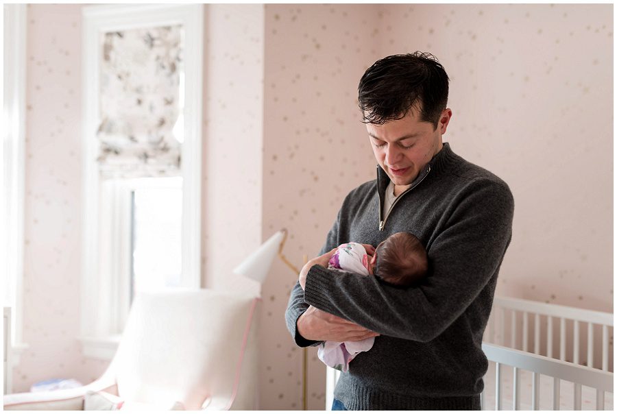 Father embraces his newborn daughter in front of the crib in her nursery.