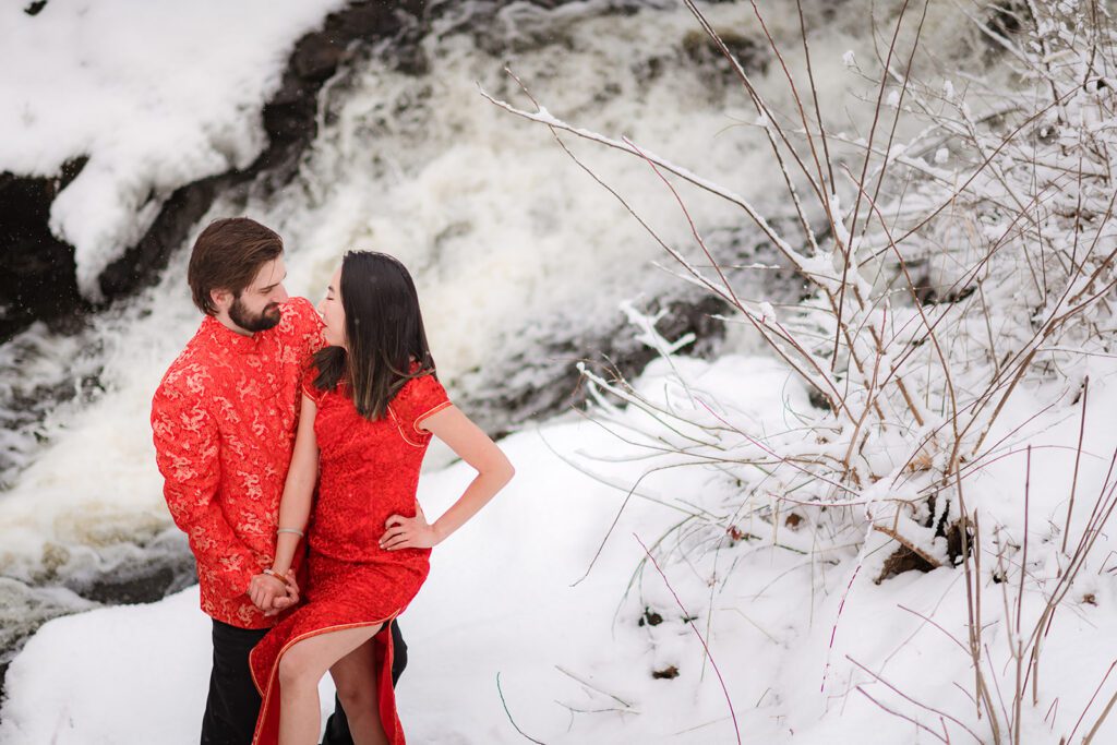 Engagement photos near Davis Square, Somerville MA — couple holding hands in nature with a river in the background taken during the winter with snow-covered ground. Couple is celebrating tradition with qipaos.